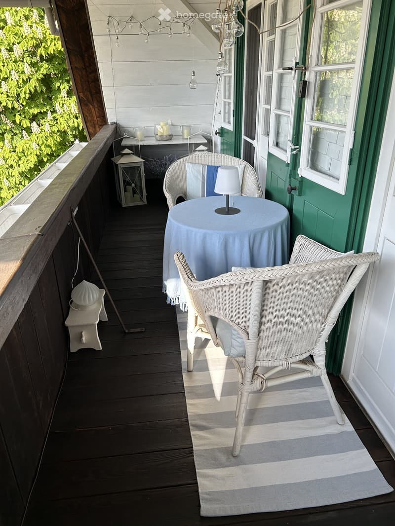 Cozy balcony with wooden decking, white wicker furniture, small table with lamp, lantern, and candles.
