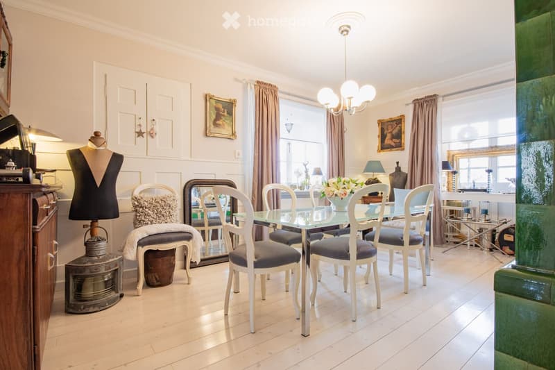 Elegant dining room with a glass dining table, six white chairs with blue cushions, a chandelier, and decorative items including a mannequin, mirror, and picture frames.