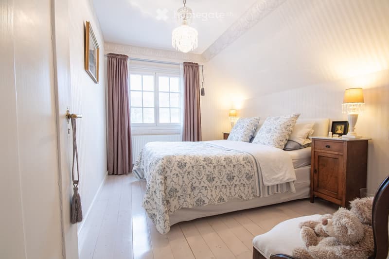 Cozy bedroom with a large bed, patterned bedspread, nightstand with lamp, wooden chair with a stuffed animal, and decorative picture frame on the wall.
