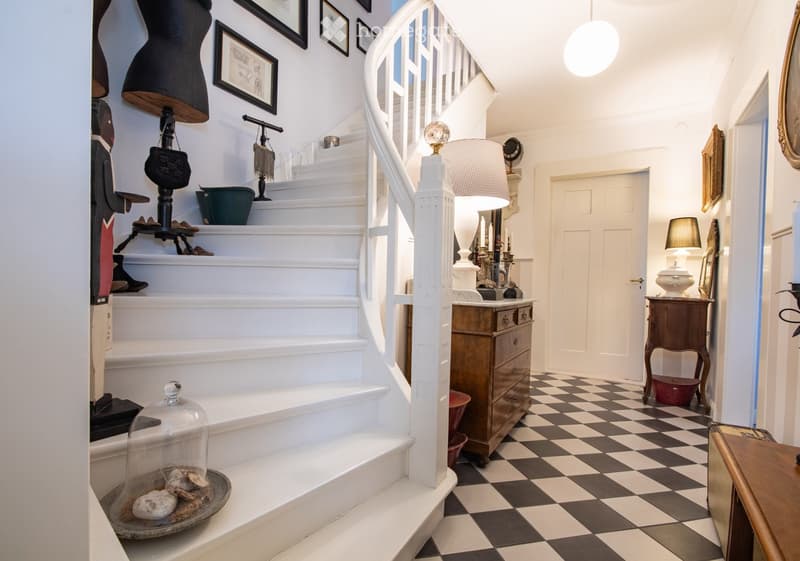 Elegant hallway with checkered black and white flooring, white staircase with decorative railings, framed artwork, and various decorative items including a glass dome with seashells, a wooden chest of drawers, and a small table with a lamp.