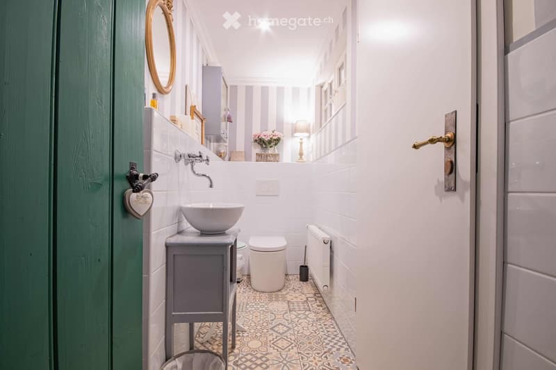 Cozy bathroom with white tiles, a sink on a grey wooden cabinet, a toilet, a mirror, a radiator, and a small table lamp on a shelf. The floor features patterned tiles, and the walls are painted white with grey stripes. A green door is visible on the left.