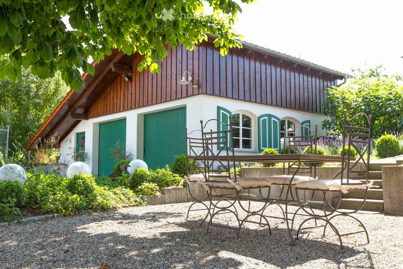 Garden area with a metal table and chairs, surrounded by greenery and a wooden house with green shutters.