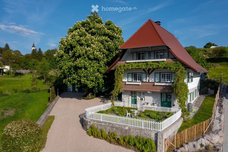 Two-story house with red-tiled roof, white walls, green shutters, and balcony. Surrounded by a well-maintained garden with a white picket fence and a large flowering tree.