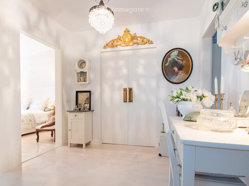 Elegant hallway with a light blue table, white chair, vase with flowers, candle holders, picture frames, and a small cabinet. A bed and nightstand are visible in the adjacent room.