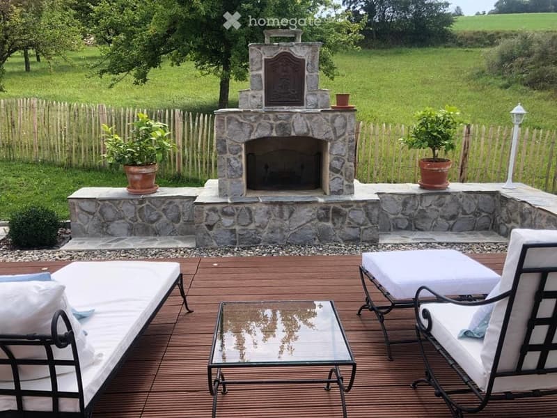 Outdoor patio with wooden decking, glass coffee table, white lounge chairs, stone fireplace, potted plants, and a white lantern post.