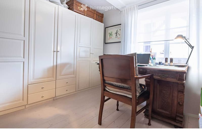 A well-lit room with a wooden chair, a wooden desk, a laptop, a lamp, a large cabinet, and a picture frame on the wall.