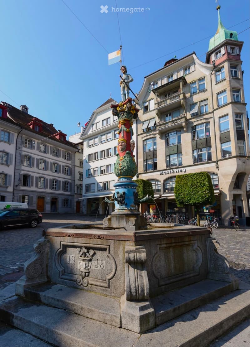 A historic square with a decorative fountain, surrounded by multi-story buildings with classic European architecture, bicycles, and a parked car.