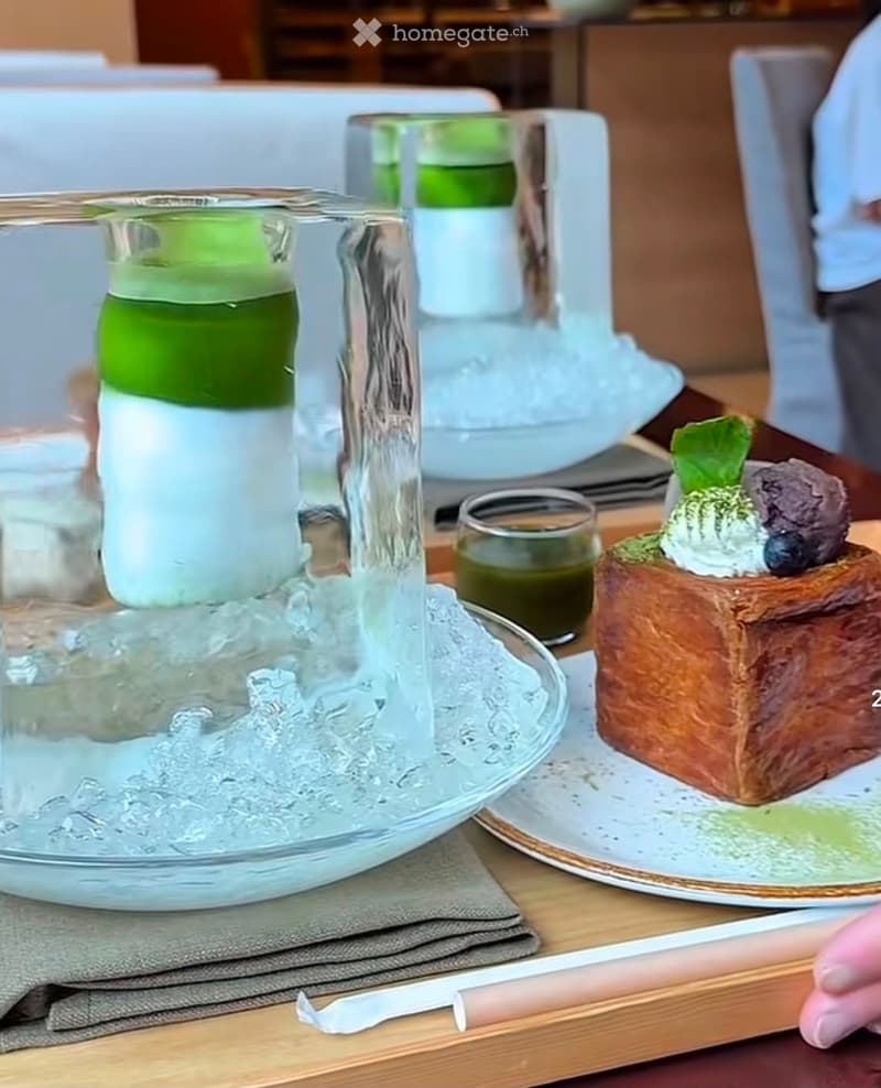A small, elegantly presented dessert on a plate, accompanied by a glass of water and a small drink in a clear glass. The dessert features a brown base, green topping, and a small dollop of cream with berries.