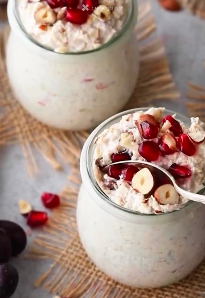 Two glass jars filled with a creamy oatmeal mixture topped with pomegranate seeds and sliced almonds, placed on a woven mat with scattered pomegranate seeds and almonds around.