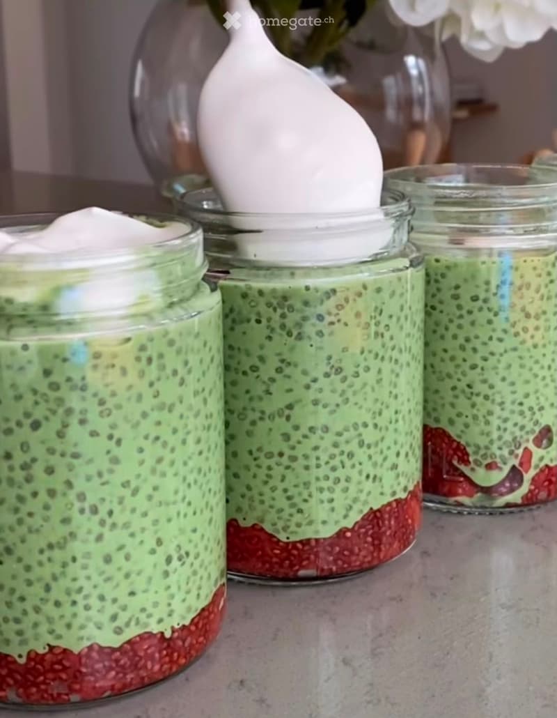 Four mason jars filled with layered green smoothie mixtures topped with whipped cream, placed on a countertop.