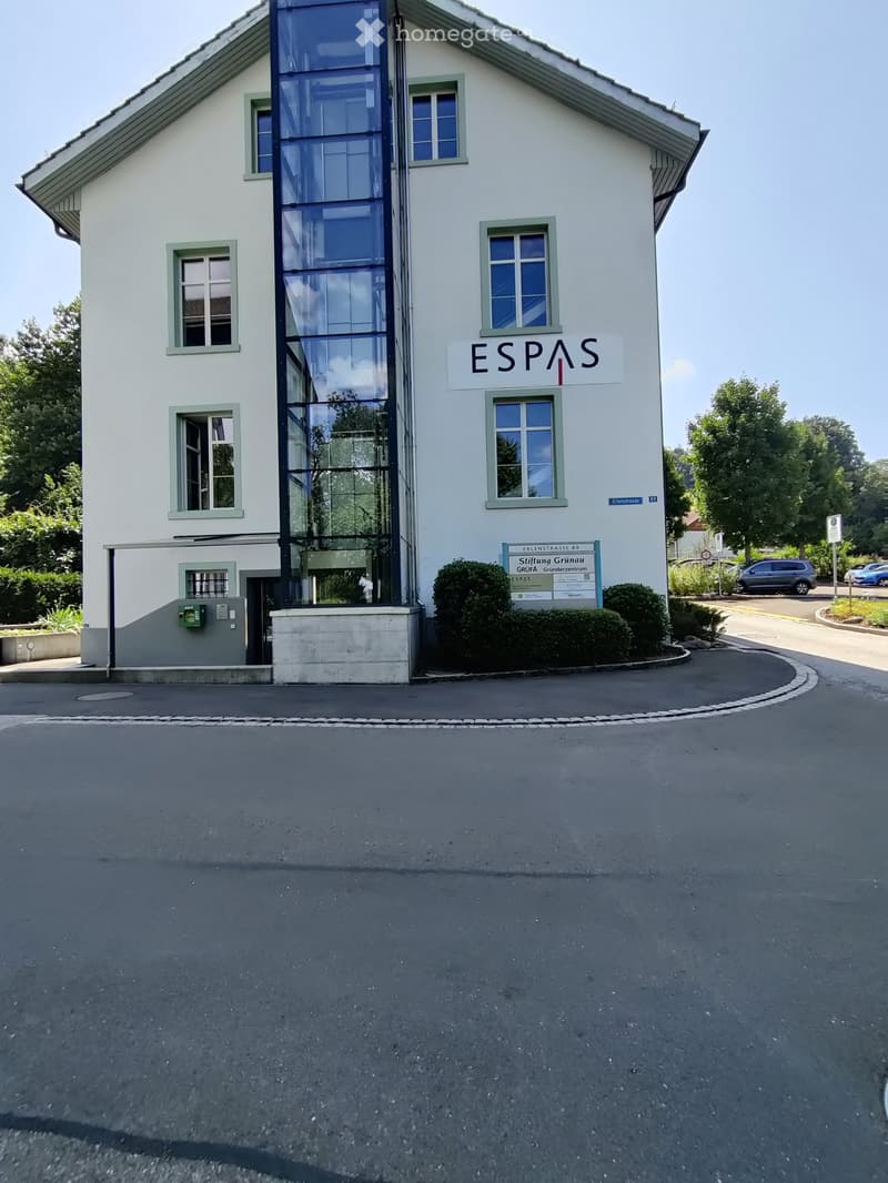 Two-story building with white facade, green-framed windows, and a modern glass elevator. Sign reads "ESPAS" and "Stiftung Grunau" with additional signage below.
