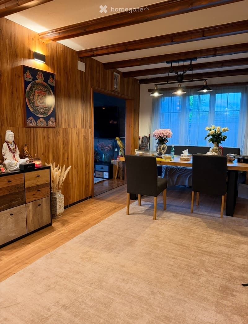 A modern living room with wooden walls, a large window, a sofa, a dining table with chairs, decorative vases, a statue, and a television in the background.