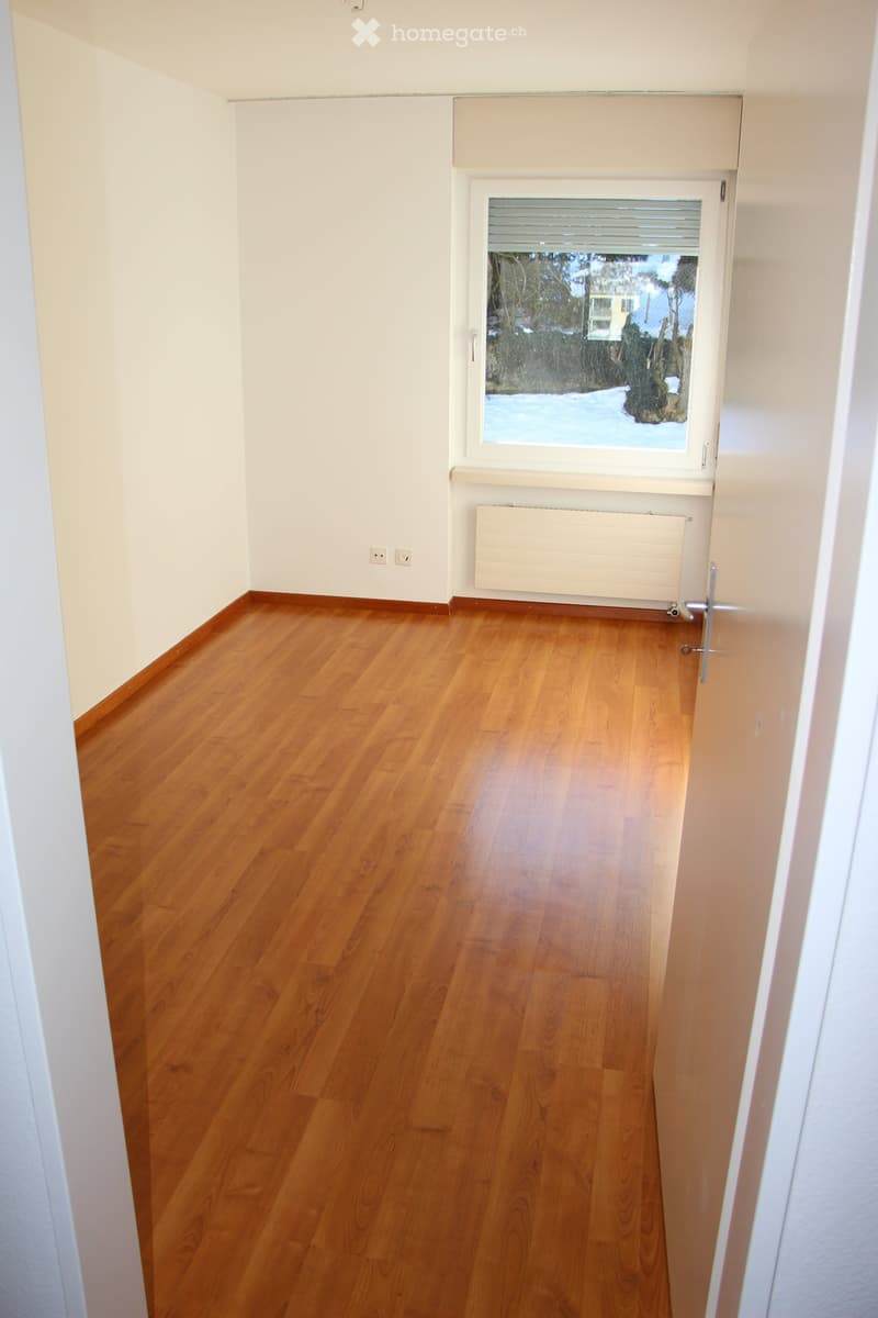Empty hallway with wooden flooring, white walls, a window with blinds, and a radiator. No furniture or personal items are present.