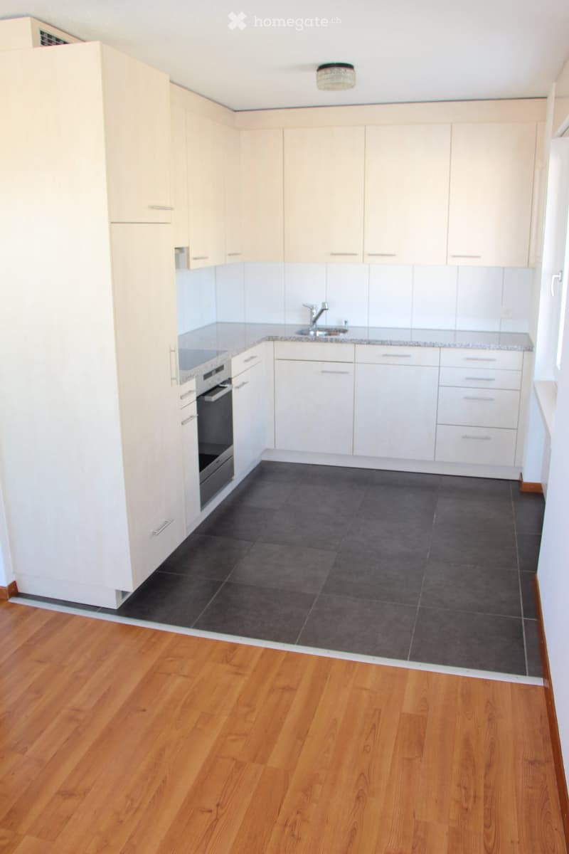 Empty kitchen with white cabinets, dark tiled floor, and wooden flooring visible in the adjacent area. No appliances or furniture are present.