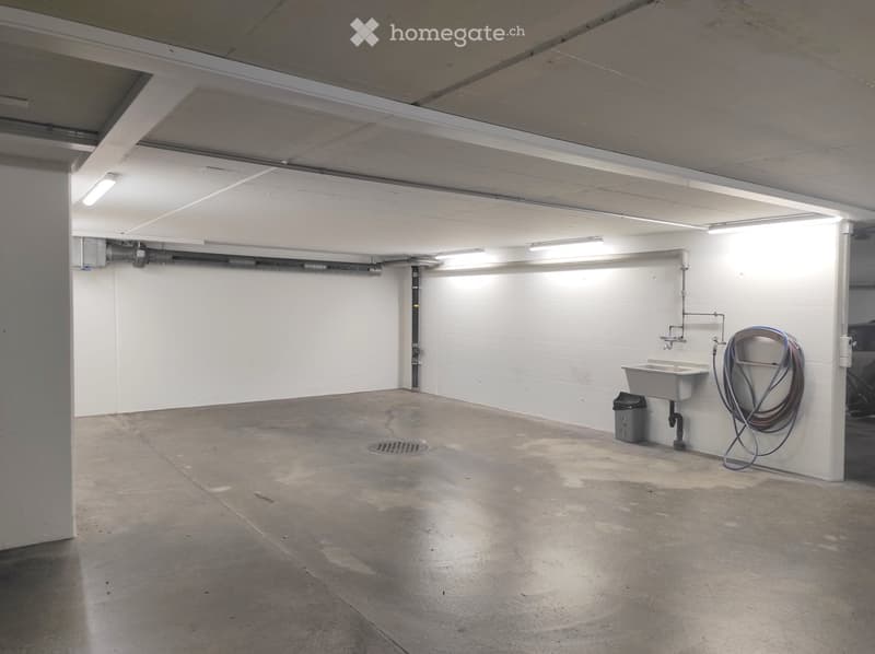 Empty concrete-walled room with a sink, faucet, and coiled hose on the wall, illuminated by overhead fluorescent lighting.