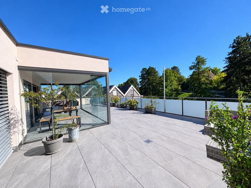 Modern balcony with grey tiled flooring, glass railing, and potted plants, featuring a small bench and a view of surrounding greenery.