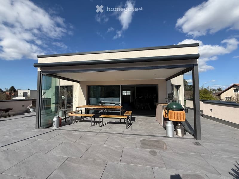 Outdoor dining area with a table and benches, glass walls, and a grill.