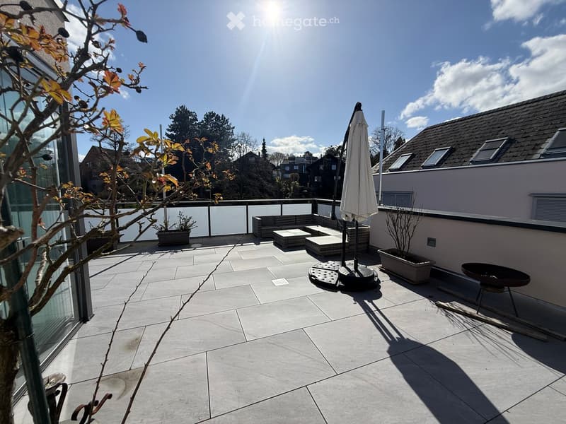 A spacious terrace with a glass railing, a closed umbrella, a sofa, a table, and potted plants.