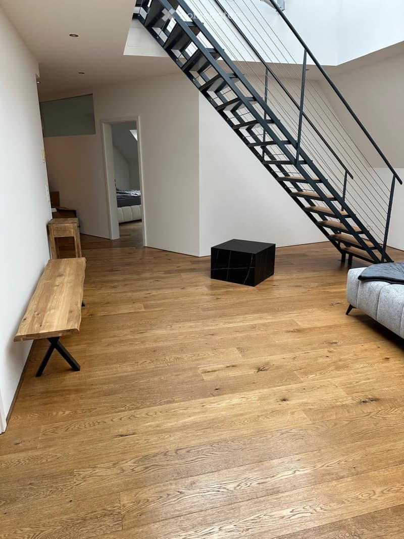 Modern living room with wooden flooring, a grey sofa, a wooden bench, and a black stool. A staircase leads to an upper level.