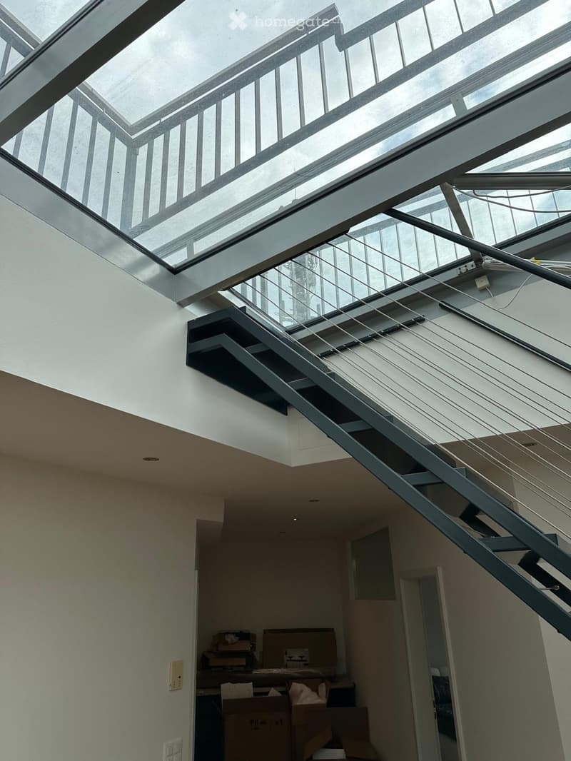 Modern interior staircase with glass ceiling and metal railings, leading to an open area with visible storage boxes and a doorway.