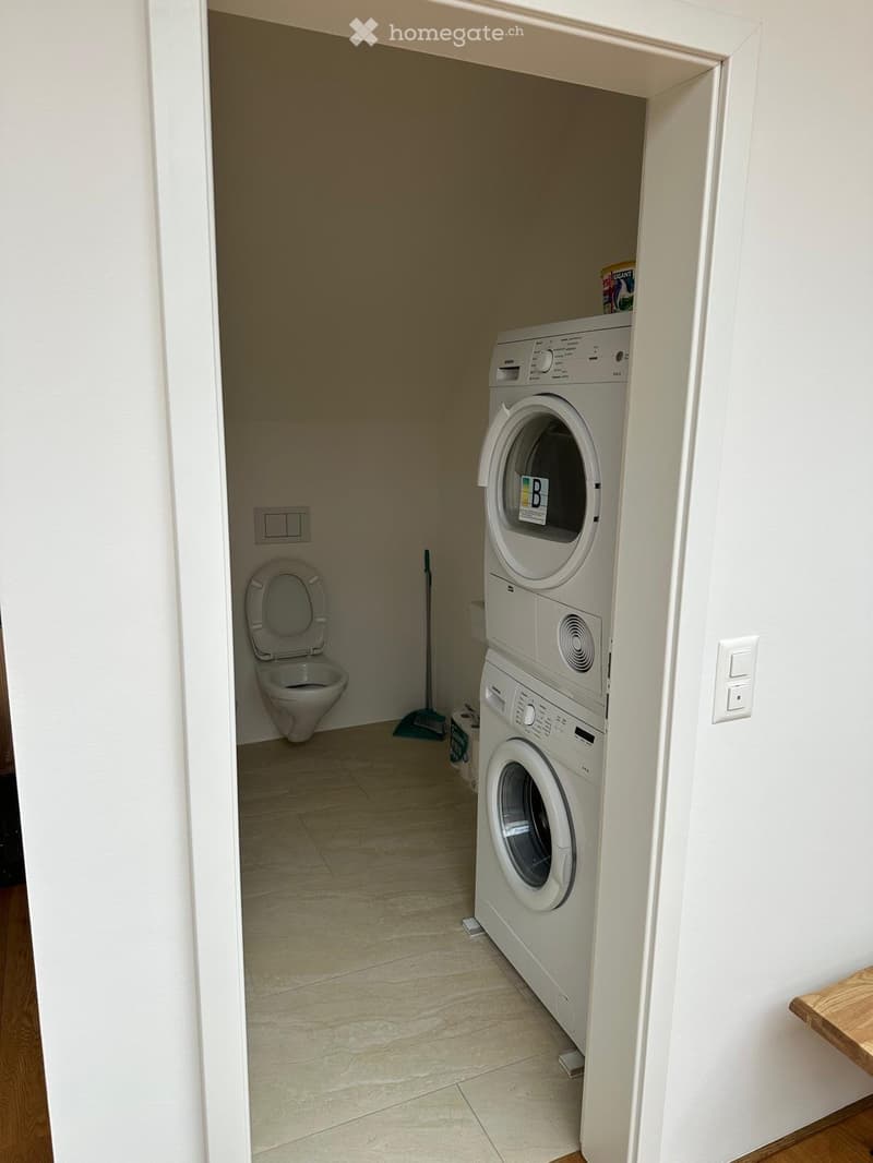 Compact bathroom with a toilet, washing machine, dryer, and a mop leaning against the wall.