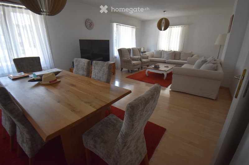 Bright living and dining area with a wooden dining table, grey upholstered chairs, a sofa, armchairs, a coffee table, and a television. The space features large windows with white curtains and a red area rug.