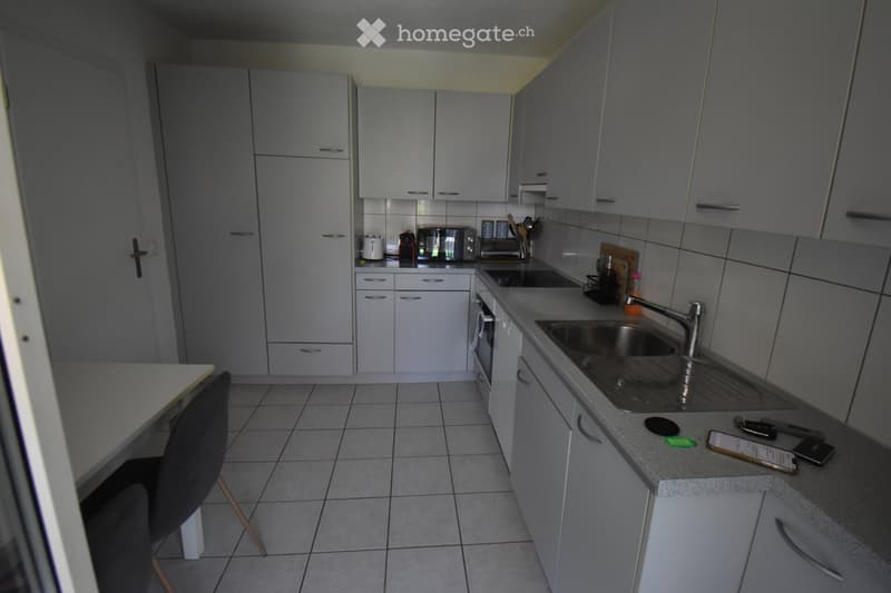 Modern kitchen with grey cabinets, tiled floor, sink, oven, microwave, toaster, and a chair near the counter.