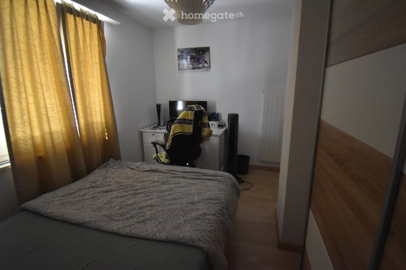A furnished bedroom with a bed, desk, chair, and a screen. The room features wooden flooring, a window with curtains, and a picture on the wall.