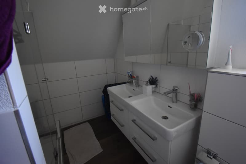 Modern bathroom with a double sink, mirrored cabinet, and tiled walls. Includes a toothbrush holder and a towel on the floor.