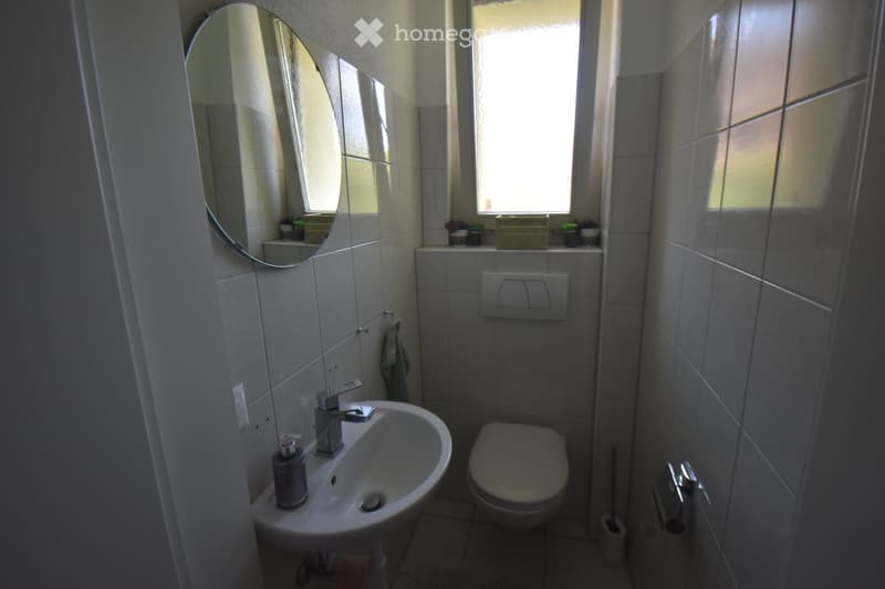 A small bathroom with a white sink, chrome faucet, toilet, and a round mirror above the sink. The walls are tiled in light gray, and a window allows natural light to enter.