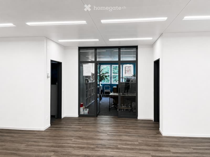 Empty room with wooden flooring, white walls, and recessed lighting. A glass door leads to an adjacent office space with desks, chairs, and office equipment.