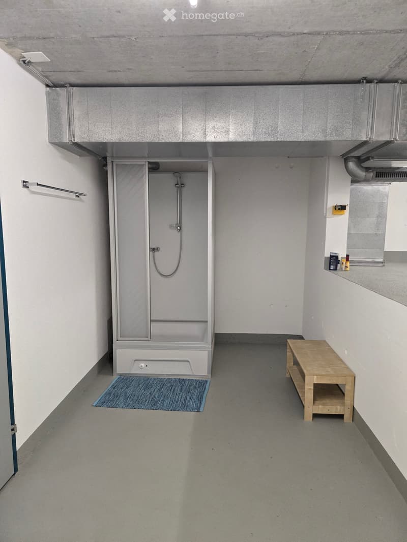 Minimalist room with a shower stall, a small wooden table, and a bench, featuring a concrete floor and white walls.