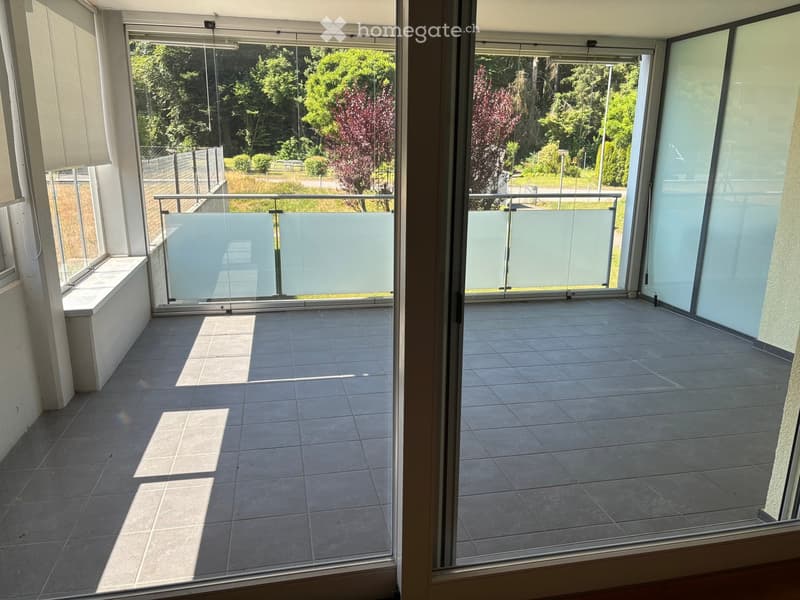 Spacious balcony with tiled flooring, large glass sliding doors, and a view of a green park area with trees and a pathway.
