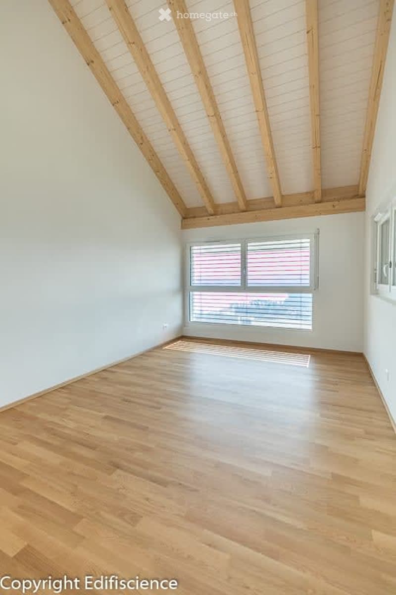 Empty room with wooden flooring and exposed wooden beams on the ceiling. Large window with blinds allows natural light to enter.