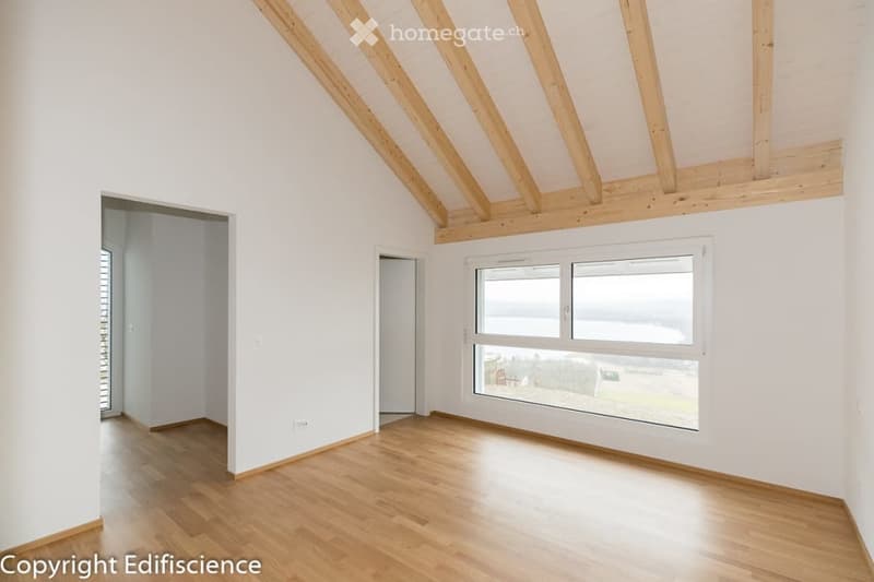 Empty room with wooden flooring, white walls, and large windows offering a view. The ceiling features exposed wooden beams.