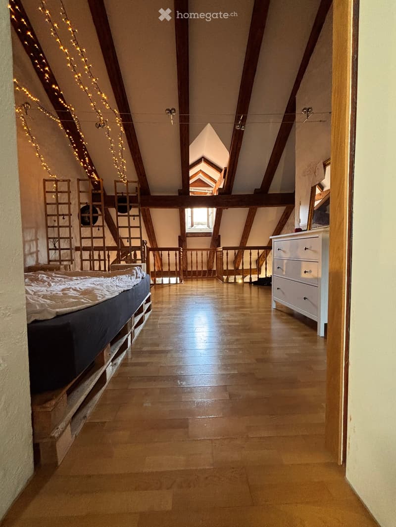 Cozy attic bedroom with wooden beams, exposed wooden floor, and a bed on the left side. Features a small balcony area with wooden railings and string lights.