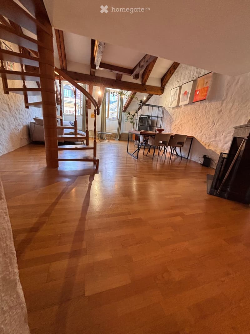 Spacious, well-lit room with wooden flooring, a curved staircase, a dining area with a table and chairs, a sofa, shelves, and artwork on the walls.