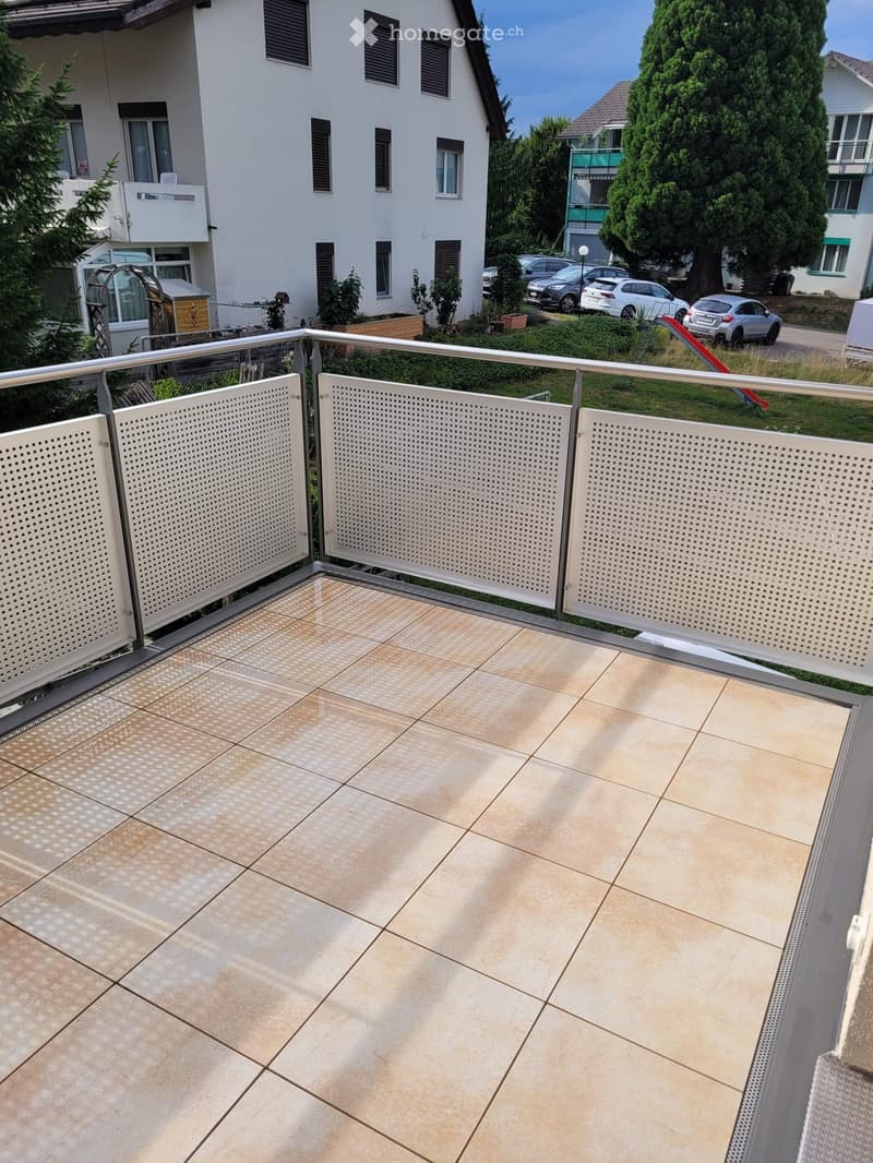 Balcony with beige tiled flooring, metal railing, and a view of a residential area with parked cars and greenery.