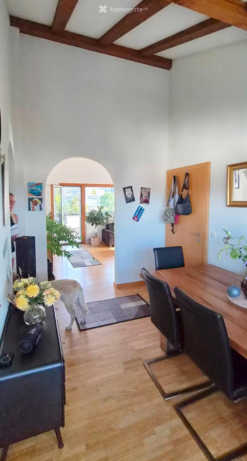 Bright hallway with wooden flooring, a dining area with a table and chairs, a vase with flowers, and a dog walking through. There is a sofa visible in the adjacent room.