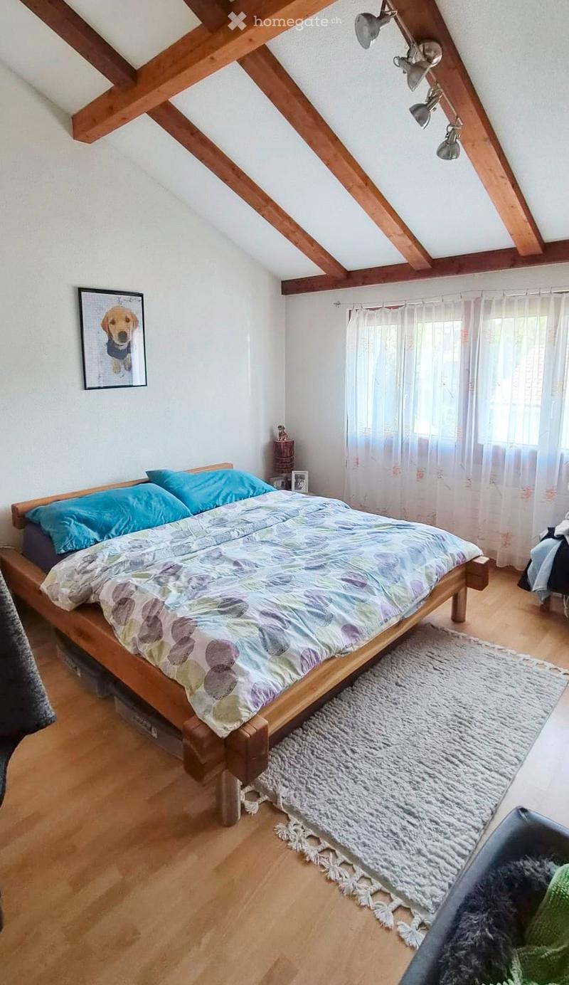 Cozy attic bedroom with wooden slanted ceilings, a wooden bed with a colorful duvet, a nightstand, a framed picture, and a white curtained window.
