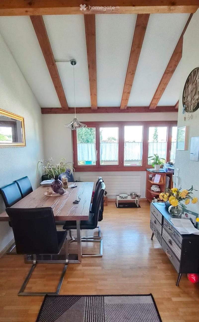 Bright dining area with wooden table, black chairs, pendant light, and decorative vases. Features large windows, wooden beams, and a small cabinet with books and flowers.