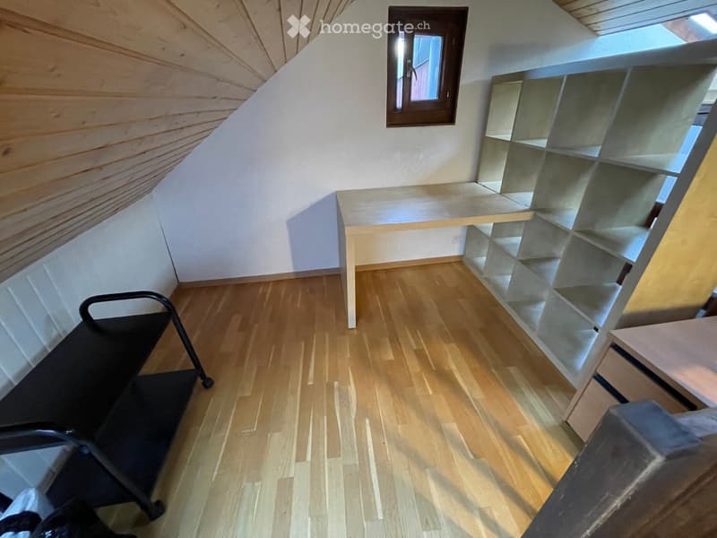 A small, modern room with wooden flooring, white walls, and a slanted ceiling. Features include a small table, a sleek shelf unit, and a black rolling cart.