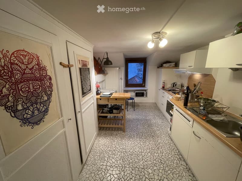 The image shows a compact kitchen with white cabinets, a wooden countertop, and a mosaic tile floor. Visible appliances include a stove, microwave, and sink. A small table with a stool is positioned near the window, and various kitchen items are scattered
