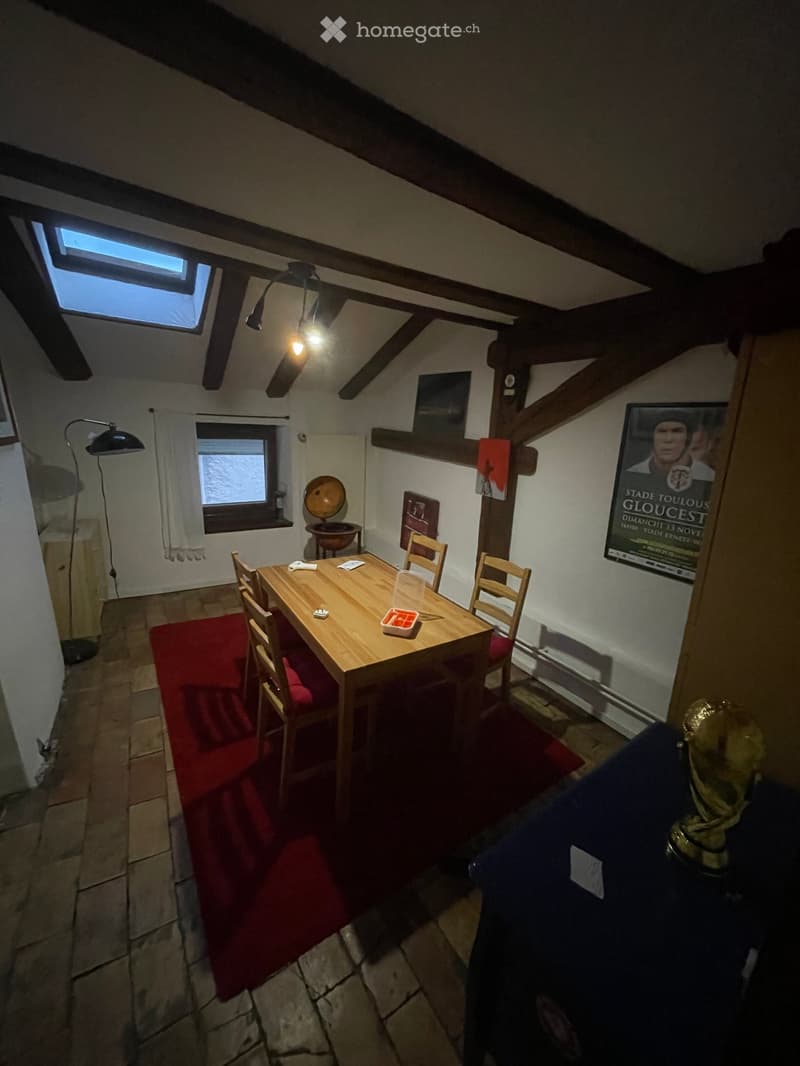 Cozy attic dining area with wooden dining table, chairs, floor lamp, and exposed wooden beams. Features a red rug, posters, and a globe on a shelf.