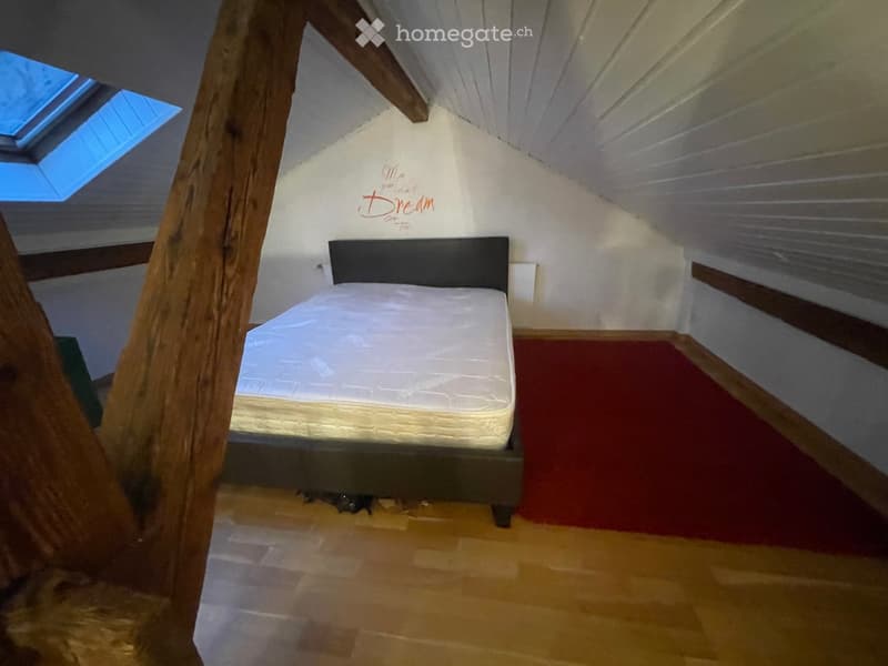 Cozy attic bedroom with wooden beams, skylight windows, and a single bed with a white mattress and dark headboard.