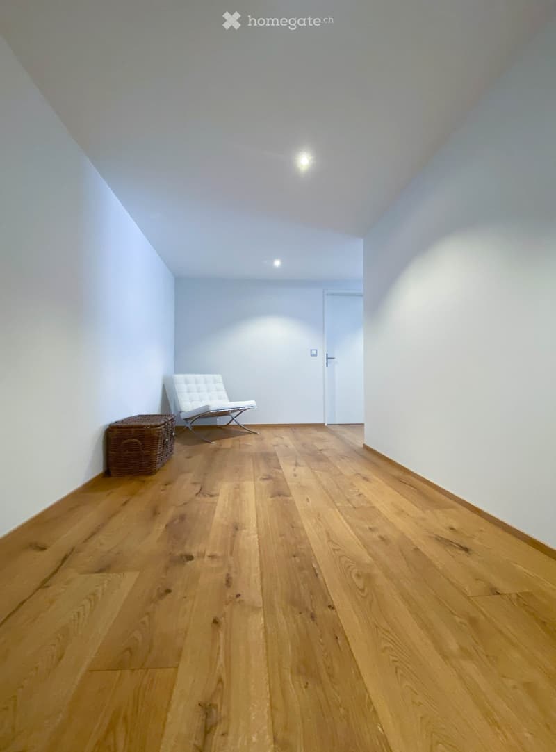 A small, well-lit room with wooden flooring, white walls, and a slanted ceiling. Features include a white modern chair and a wicker basket near the wall.
