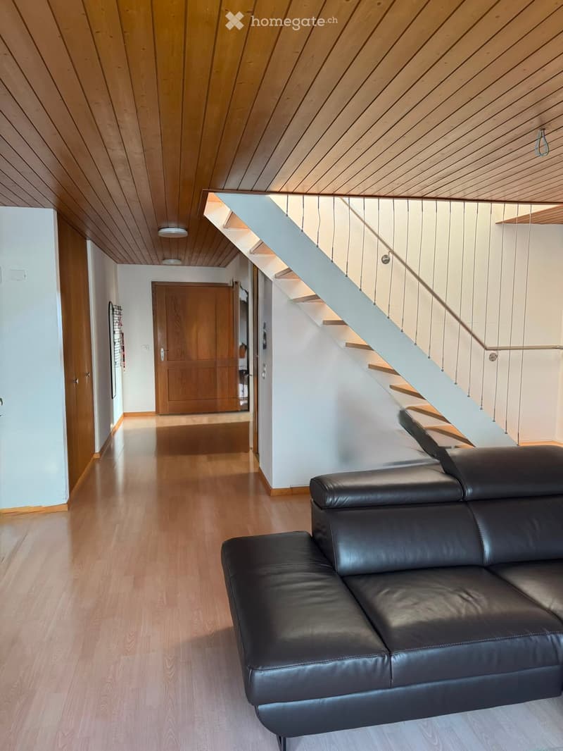 Modern hallway with wooden flooring, a sleek staircase with a glass railing, and a black leather sofa placed near the staircase.