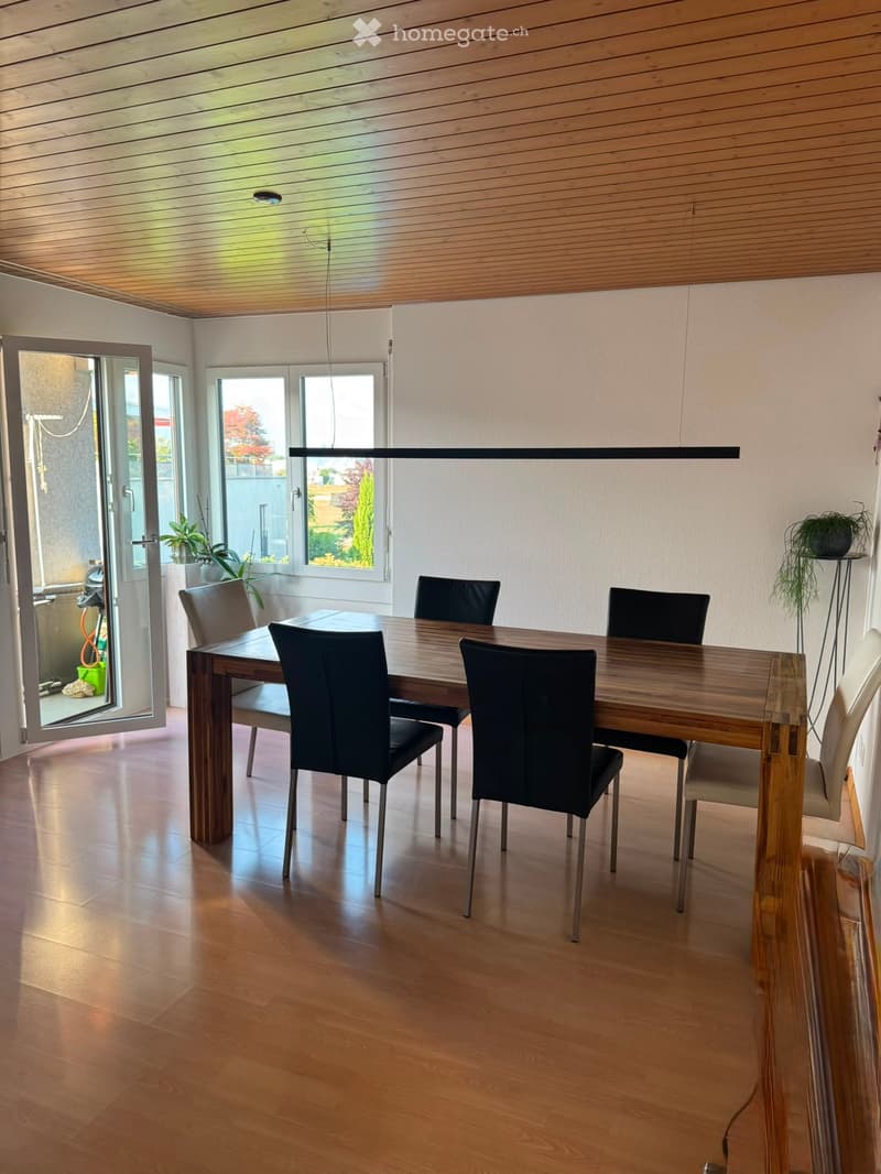 Dining room with wooden floor, modern wooden dining table, black chairs, pendant light, large windows, and a view of the garden.