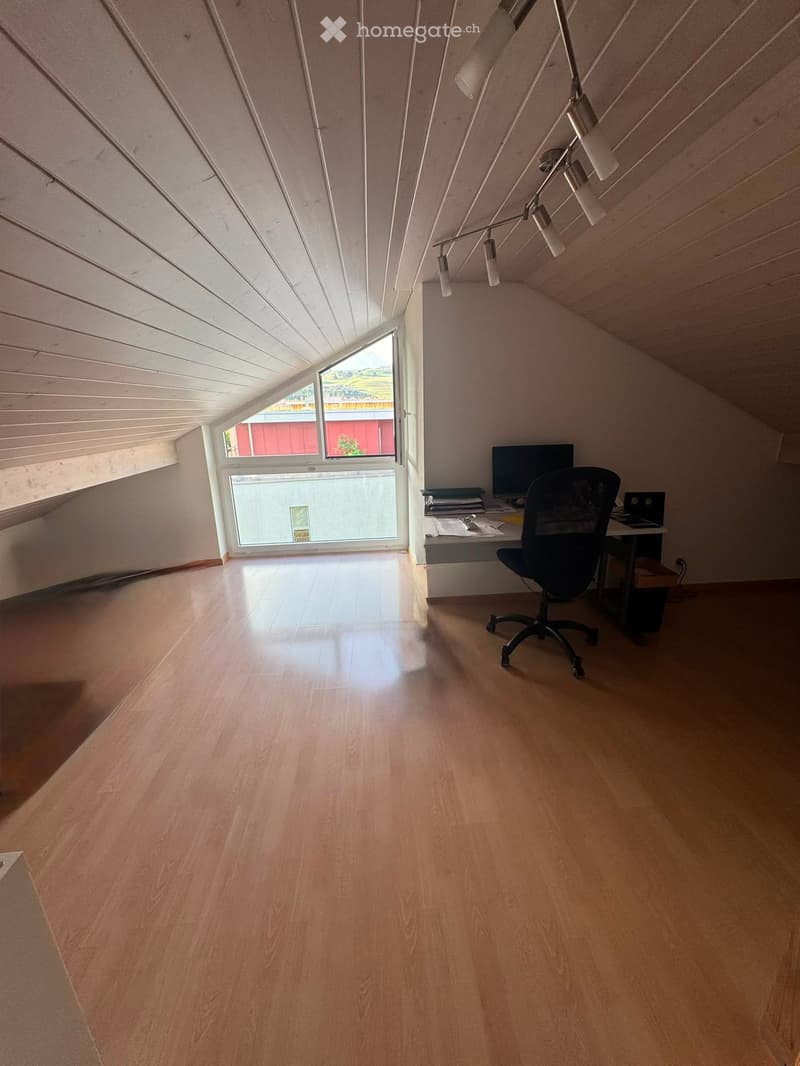 Bright attic room with wooden floors, slanted ceilings, and large windows offering a view. Contains a desk with a computer, chair, and laptop.