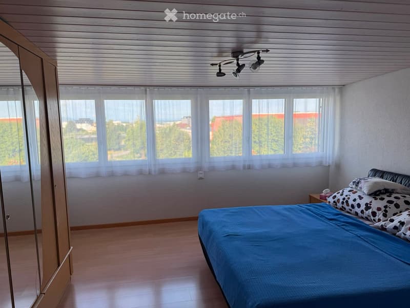 Bright bedroom with wooden flooring, large windows with white curtains, a bed with blue bedding and patterned pillows, a nightstand, and a mirrored wardrobe.